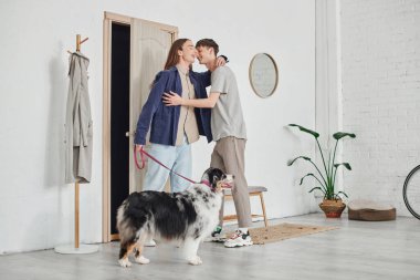 Mutlu eşcinsel çift, gündelik giysiler içinde koridorda, askılığın yanında durup birbirlerine sarılıyorlar ve modern apartmanda birlikte gülümserken Avustralya çoban köpeğinin yanında tasma tutuyorlar.