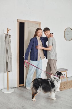 happy gay couple in casual outfits standing and hugging each other in hallway next to coat rack and holding leash near Australian shepherd dog and smiling together in modern apartment clipart