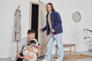 Mutlu eşcinsel adam gülümserken, modern koridorda kapıyı açarken uzun saçlı neşeli erkek arkadaşının yanında sevimli Avustralyalı çoban köpeğine sarılıyor. 