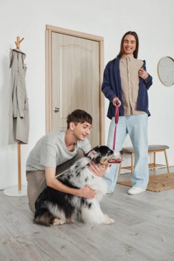 Neşeli eşcinsel adam gülümserken, modern koridorda kapıya yakın dururken uzun saçlı neşeli erkek arkadaşının yanında sevimli Avustralyalı çoban köpeğine sarılıyor. 