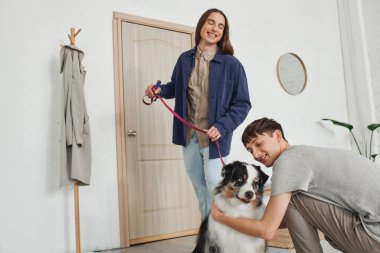 Mutlu gay adam gülümserken Avustralya çoban köpeğine sarılıyor. Uzun saçlı, neşeli erkek arkadaşının yanında. Modern koridorda kapı ve askıların yanında duruyor. 