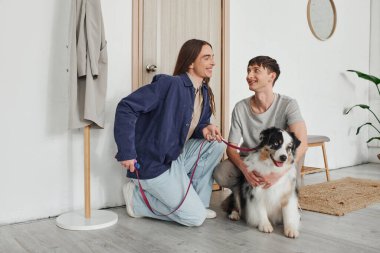 Neşeli Lgbt çifti, gündelik giysiler içinde gülümserken birlikte diz çökerken ve Avustralya çoban köpeğine sarılırken modern apartmanın koridorunda kapı ve askılık yanında. 