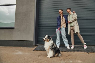 cheerful gay man with pigtails holding leash of Australian shepherd dog and hugging smiling boyfriend in casual outfit while standing together near garage door outside on street clipart