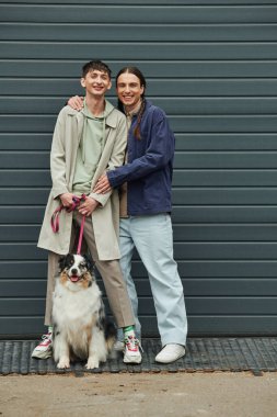 Saçları örgülü, neşeli, sırıtan, sırıtan, erkek arkadaşı dövmeli, tasmalı, çoban köpeği tasmalı ve caddede garaj kapısının yanında duran neşeli bir gay.