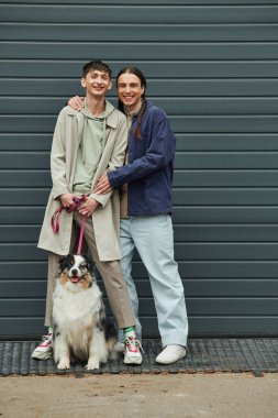 cheerful gay man with pigtails hugging smiling tattooed boyfriend in casual outfit holding leash of Australian shepherd dog and standing next to near garage door outside on street clipart