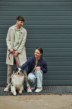cheerful gay man with pigtails cuddling Australian shepherd dog next to smiling boyfriend in coat holding leash near garage door outside on street clipart