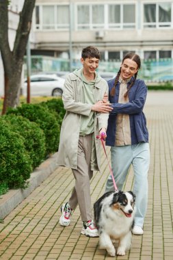 Avustralya çoban köpeğinin tasmasını tutan neşeli eşcinseller birlikte yürürken yeşil çalıların ve kentsel caddedeki modern binanın yanında gülümsüyorlar. 