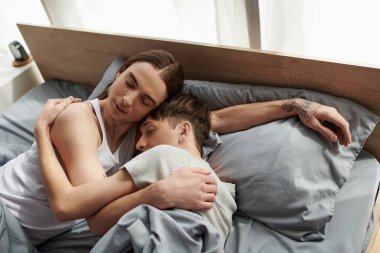 Uzun saçlı, kapalı gözlü, rahat yastığın üzerinde, battaniyenin altında yatan ve modern yatak odasında erkek arkadaşına sarılan genç bir gay.