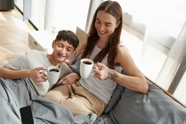 スマートフォンの近くでコーヒーカップを持ちながら 朝の快適なベッドの上で休んでいるときの笑顔のゲイカップルの高い角度ビュー — ストック写真