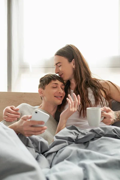 Long haired and tattooed gay man holding coffee cup and kissing smiling boyfriend in pajama while resting with smartphone in hand in modern bedroom in morning  time