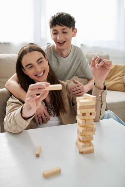 Позитивный гей в повседневной одежде обнимает татуировку и длинноволосый бойфренд держа часть размытых деревянных блоков игры на столе в гостиной дома 