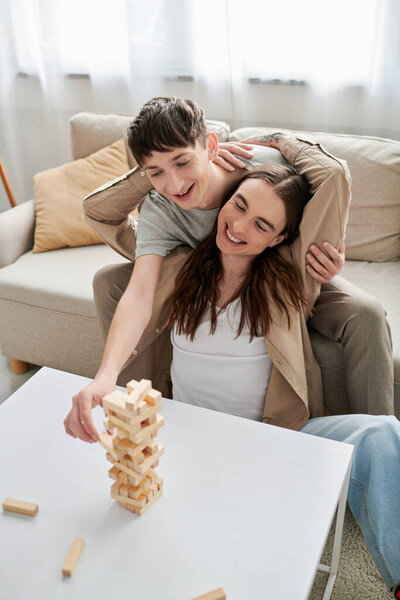 Позитивный и длинноволосый гомосексуалист обнимает бойфренда, играя в деревянные блоки на столе возле удобного дивана в гостиной 