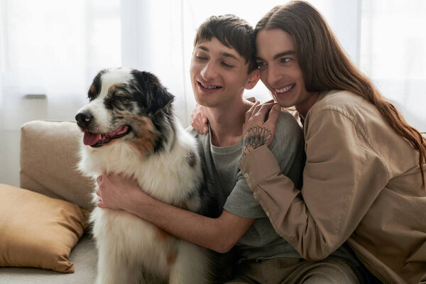 Positive long haired and tattooed homosexual man in shirt hugging young boyfriend and looking at furry Australian shepherd dog on comfortable couch at home 