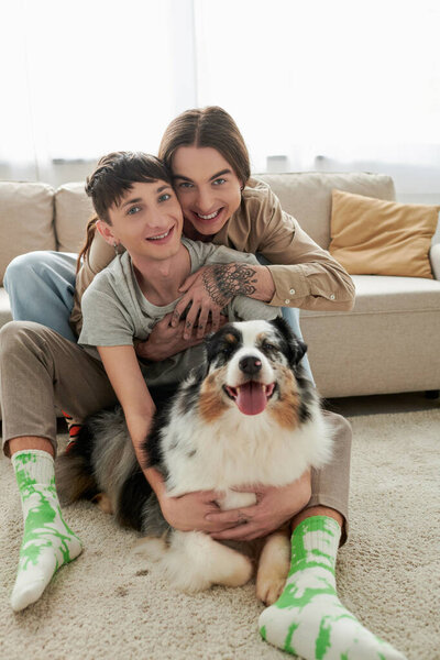 Cheerful and young lgbt couple in casual clothes and socks hugging and looking at camera near furry Australian shepherd dog lying on carpet on floor in modern living room at home 
