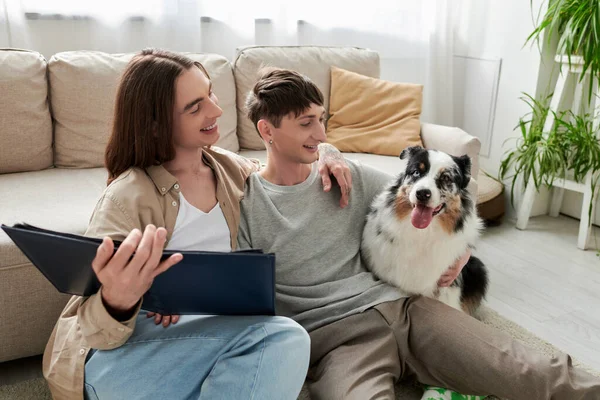 年轻快乐的同性伴侣一边抱着相册一边拥抱 在家里的现代客厅里看着地板上那只友善的澳大利亚牧羊犬 — 图库照片