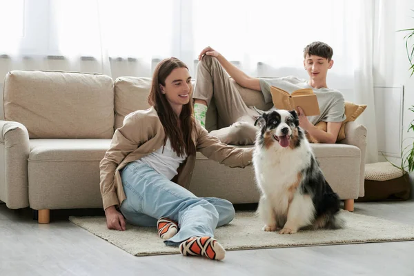 stock image happy gay man reading book and lying on comfortable couch while his boyfriend with long hair playing with Australian shepherd dog during his free time at home 