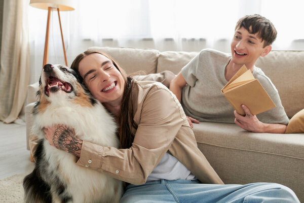 happy gay man with tattoo smiling while hugging Australian shepherd dog next to cheerful gay man holding book and resting on comfortable couch in living room 