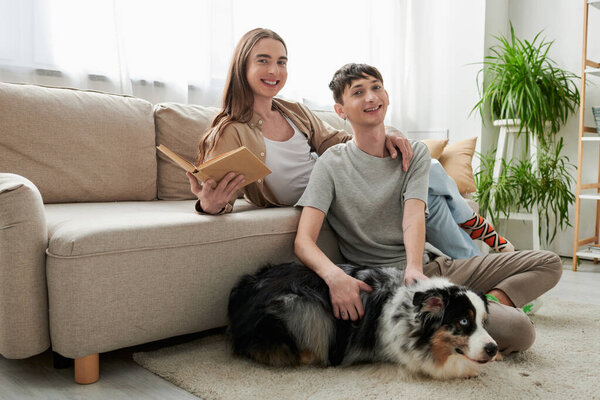 cheerful gay man sitting on carpet and cuddling Australian shepherd dog next to happy partner with long hair holding book and looking at camera in modern living room 