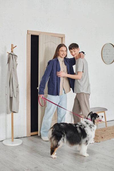 happy gay couple in casual outfits standing and hugging each other in hallway next to coat rack and holding leash near Australian shepherd dog and smiling together in modern apartment