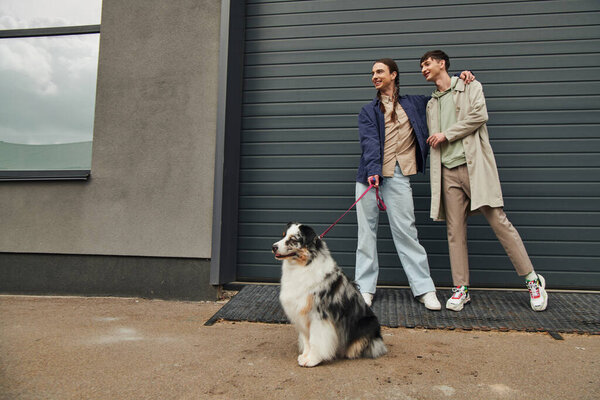 cheerful gay man with pigtails holding leash of Australian shepherd dog and hugging smiling boyfriend in casual outfit while standing together near garage door outside on street