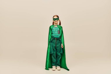 Yüzünde pelerin ve maskeyle yeşil süper kahraman kostümü içinde gülümseyen, pantolon ve tişört giyen ve gri arka planda çocuk koruma günü tatilini kutlayan bir kız. 