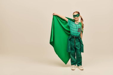 happy kid in superhero costume and mask on face holding green cloak, wearing pants and t-shirt and standing while celebrating Child protection day holiday on grey background  clipart
