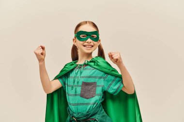 Gri arka planda Dünya Çocuk Koruma Günü tatilini kutlarken yüzünde pelerin ve maskeyle gülümseyen ve yumruklarıyla ayakta duran yeşil süper kahraman kostümlü güçlü bir kız. 