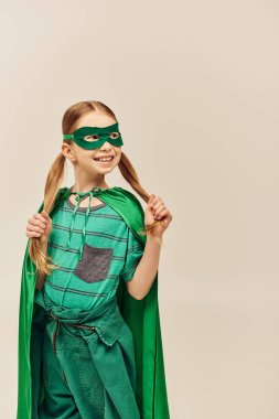 Yeşil süper kahraman kostümlü mutlu kız yüzünde maske ve pelerin, saçında ikiz kuyruk ile gri arka planda Dünya Çocuk Koruma Günü 'nü kutlarken saçına dokunuyor. 