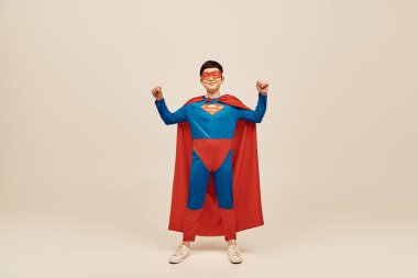 happy asian boy in red and blue superhero costume with cloak and mask on face showing strength gesture while celebrating Happy children's day on grey background  clipart