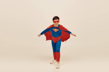 courageous asian boy in red and blue superhero costume with cloak and mask on face celebrating International Day for Protection of Children on grey background  clipart