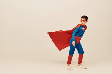 full length of courageous asian boy in superhero costume with cloak and mask looking away and standing against wind during World Child protection day on grey background  clipart