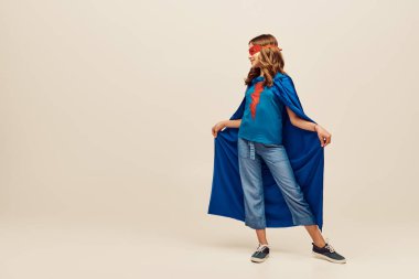 Süper kahraman kostümlü mutlu kız ve yüzünde kırmızı maske, elinde mavi pelerin, gri arka planda kot pantolon ve tişört, Uluslararası Çocukları Koruma Günü konsepti 