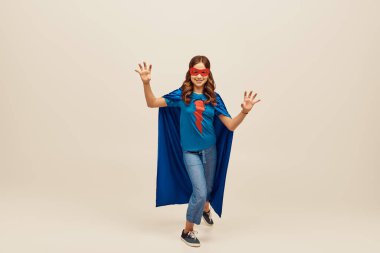 joyful girl in superhero costume with blue cloak and red mask on face, showing hand gesture, standing in denim jeans and t-shirt while celebrating Child protection day holiday on grey background  clipart