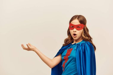 Mavi pelerinli, kırmızı maskeli, şok olmuş süper kahraman kız elini uzatarak stüdyoda gri arka planda Uluslararası Çocuk Koruma Günü 'nde ağzını açtı. 