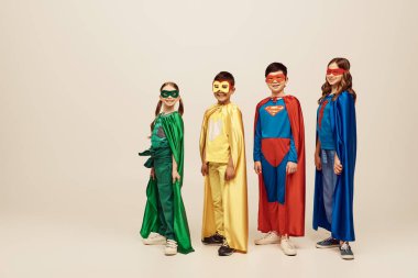 Renkli süper kahraman kostümlü mutlu ırklar arası çocuklar gri arka planda, stüdyoda, Dünya Çocuk Koruma Günü konseptinde bir arada duruyorlar. 