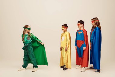 happy interracial preteen kids in colorful superhero costumes looking at girl standing in green cloak and mask while celebrating Child protection day holiday on grey background in studio  clipart