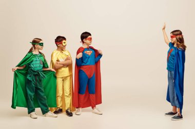 Renkli süper kahraman kostümlü mutlu ırklar arası çocuklar stüdyoda gri arka planda el sallayan kızlara bakıyorlar Uluslararası Çocukları Koruma Günü konsepti 