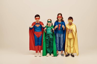 multicultural kids in colorful costumes with cloaks and masks pouting lips, looking at camera together and celebrating International children's day on grey background in studio  clipart