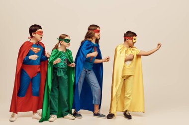 courageous multicultural kids in colorful costumes with cloaks and masks standing with clenched fists together on grey background in studio, Child Protection Day concept clipart