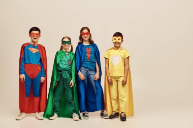 happy interracial kids in colorful costumes with cloaks and masks smiling together and looking at camera on grey background in studio, Child Protection Day concept  clipart