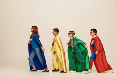side view of happy interracial kids in colorful costumes with cloaks and masks smiling and walking together on grey background in studio, Child Protection Day concept  clipart