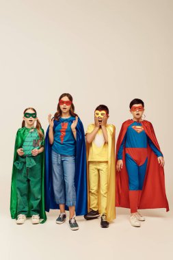 shocked interracial children in colorful superhero costumes with cloaks and masks looking at camera on grey background in studio, Child Protection Day concept  clipart
