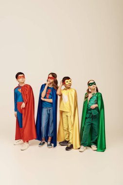 pensive multicultural kids in colorful superhero costumes with cloaks and masks scratching head and looking away while thinking on grey background in studio, children's day concept clipart