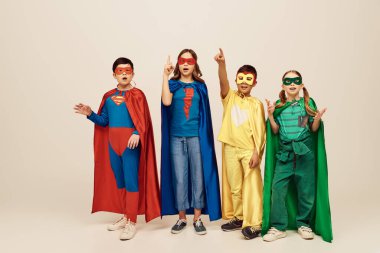 Renkli süper kahraman kostümlü çok kültürlü çocuklar fikir jestlerini parmaklarıyla gösteriyorlar ve gri arka plan hakkında konuşuyorlar stüdyoda, çocuk günü konseptinde.