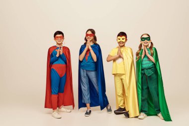 Renkli süper kahraman kostümlü mutlu çok kültürlü çocuklar dua eden ellerle ayakta duruyorlar ve gri arka planda birlikte gülüyorlar stüdyoda, Uluslararası Çocuk Günü konseptinde.