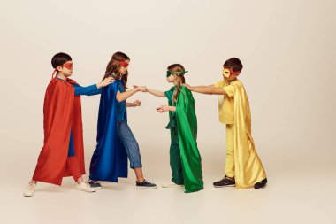 Renkli süper kahraman kostümlü, maskeli ve pelerinli ırklar arası çocukların gri arka planda stüdyoda, Uluslararası Çocuk Günü konseptinde birbirleriyle kavga etmelerinin yan görüntüsü