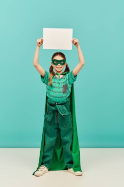 happy preteen girl in green superhero costume with cloak and mask holding blank paper above head and looking at camera while celebrating Child protection day holiday on blue background 