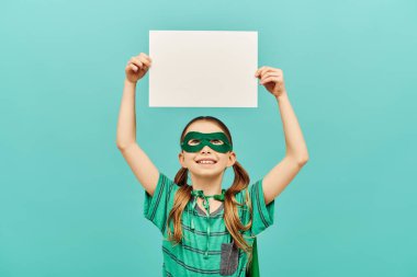 happy preteen girl in green superhero mask holding blank paper above head and looking up on blue background, World child protection day concept  clipart