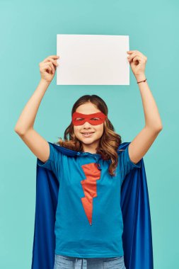 carefree preteen girl in superhero costume with cloak and red mask holding blank paper above head and looking at camera on blue background, Happy children's day concept  clipart