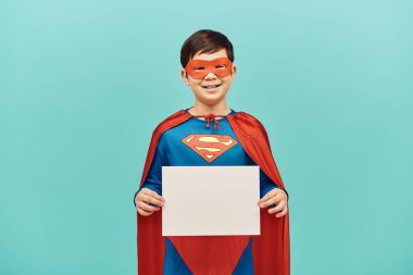 Süper kahraman kostümlü kaygısız ergen Asyalı çocuk elinde boş bir kağıtla dikilip kameraya bakıyor, dünya çocuk koruma günü konsepti. 